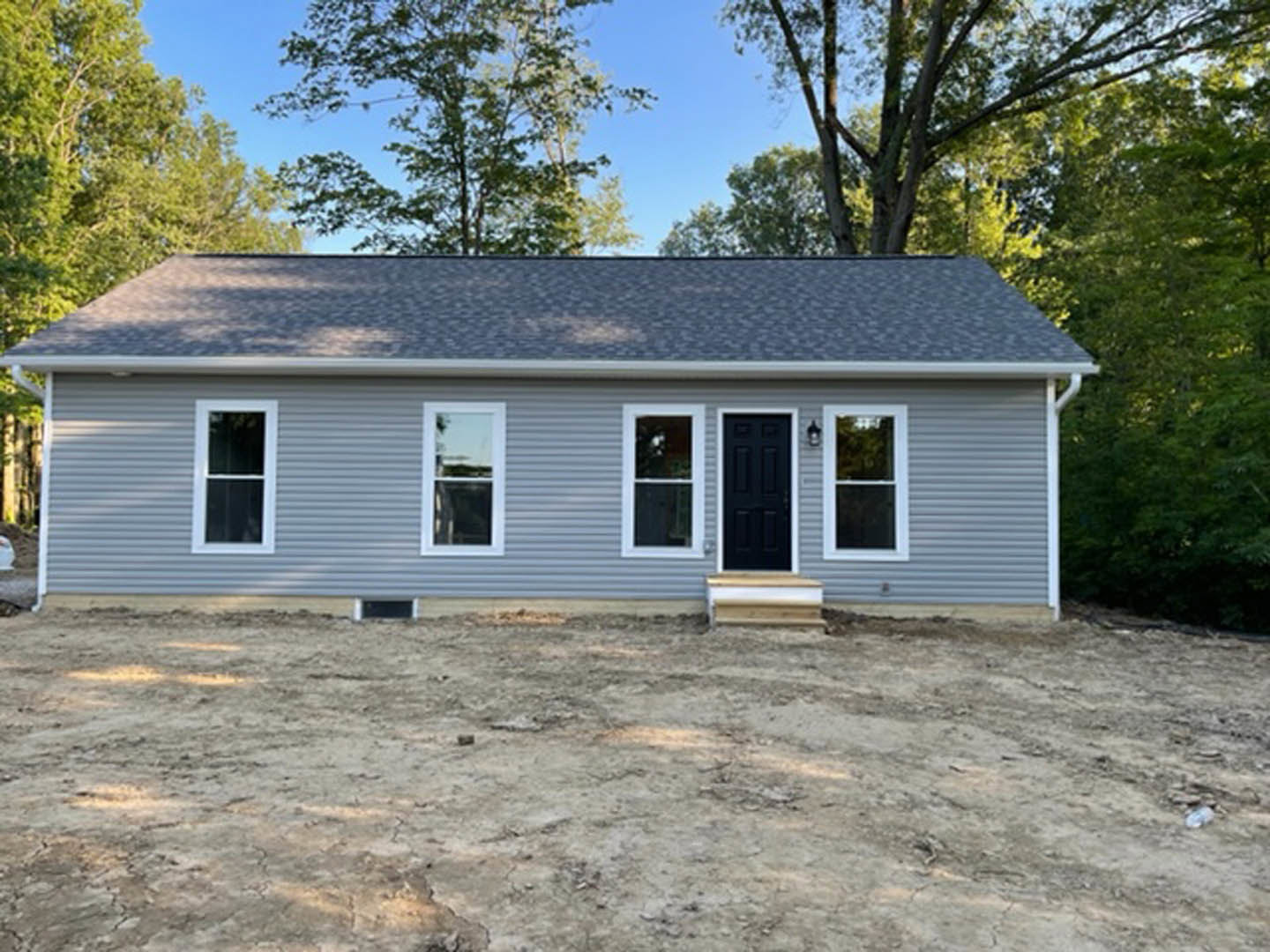 Modern home with light-colored siding, white-framed windows, metal railing along dirt yard, surrounded by mature trees.
