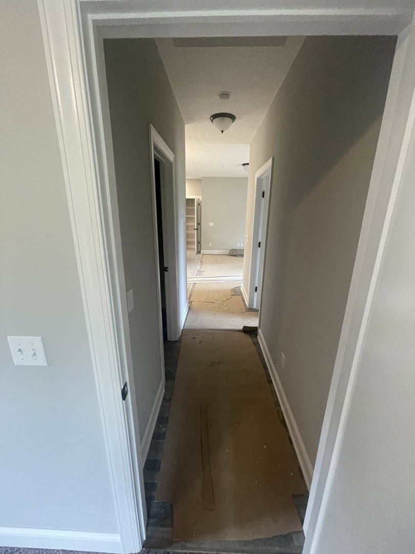 Long hallway with light brown wood flooring, white plaster walls, white ceiling, open white door with black handle, white wall outlet, simple base molding