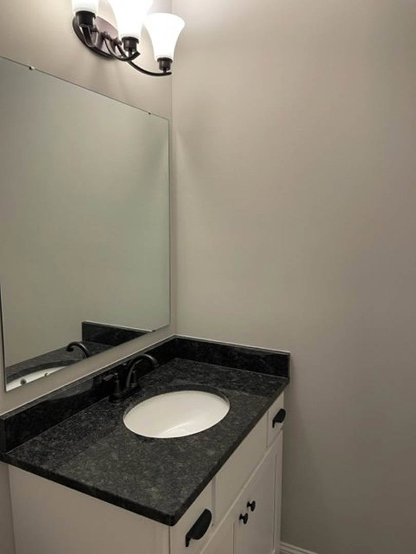 White undermount sink with chrome faucet set in black stone countertop, rectangular mirror above, white cabinetry with black handles, light fixture mounted on white tiled wall.
