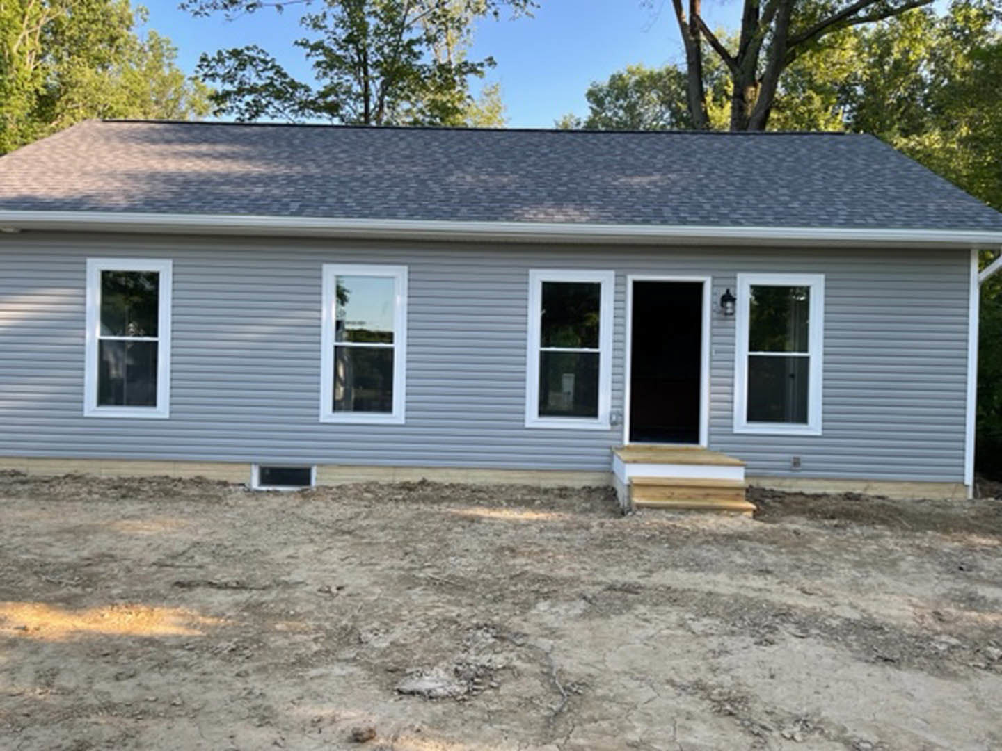 Partially built home with exposed wooden framing, open doorway, white-framed window, dirt ground, and mature trees in the background