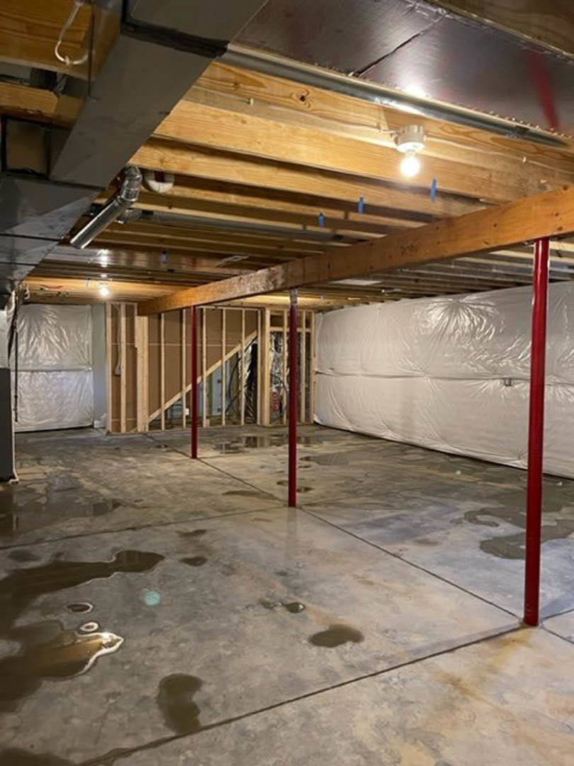 Basement with wooden plank ceiling, white painted wall, wet concrete floor with red support poles, large white plastic-wrapped object, exposed ceiling lights and pipes, close-up
