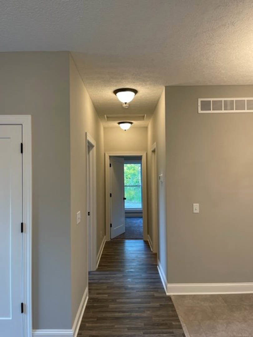 Hallway with dark wood flooring, white trim, ceiling light fixture, white door featuring a window, and close-up of a metallic door handle