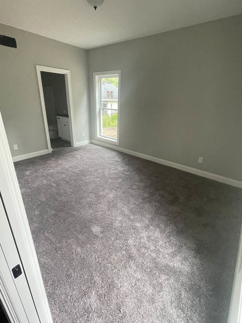 Carpeted bedroom with white walls, large window, and wooden door; natural light highlights neutral finishes.