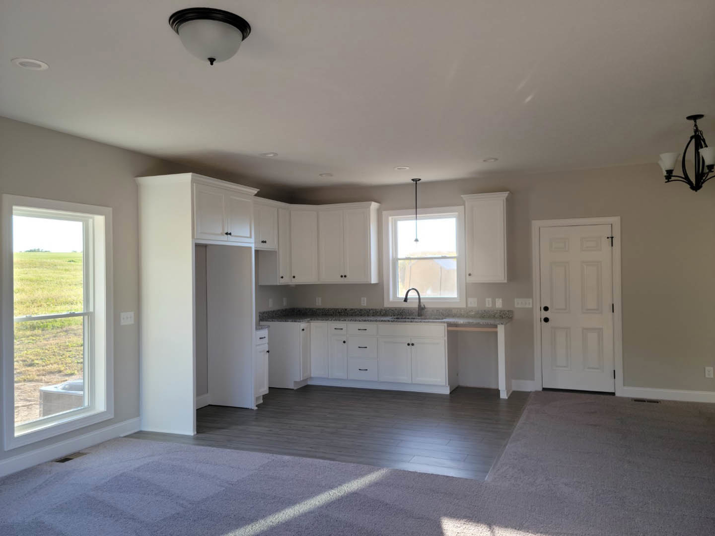 White cabinetry with black hardware, stainless steel sink beneath a window overlooking grass, light fixture on ceiling, white door with black knobs, neutral flooring