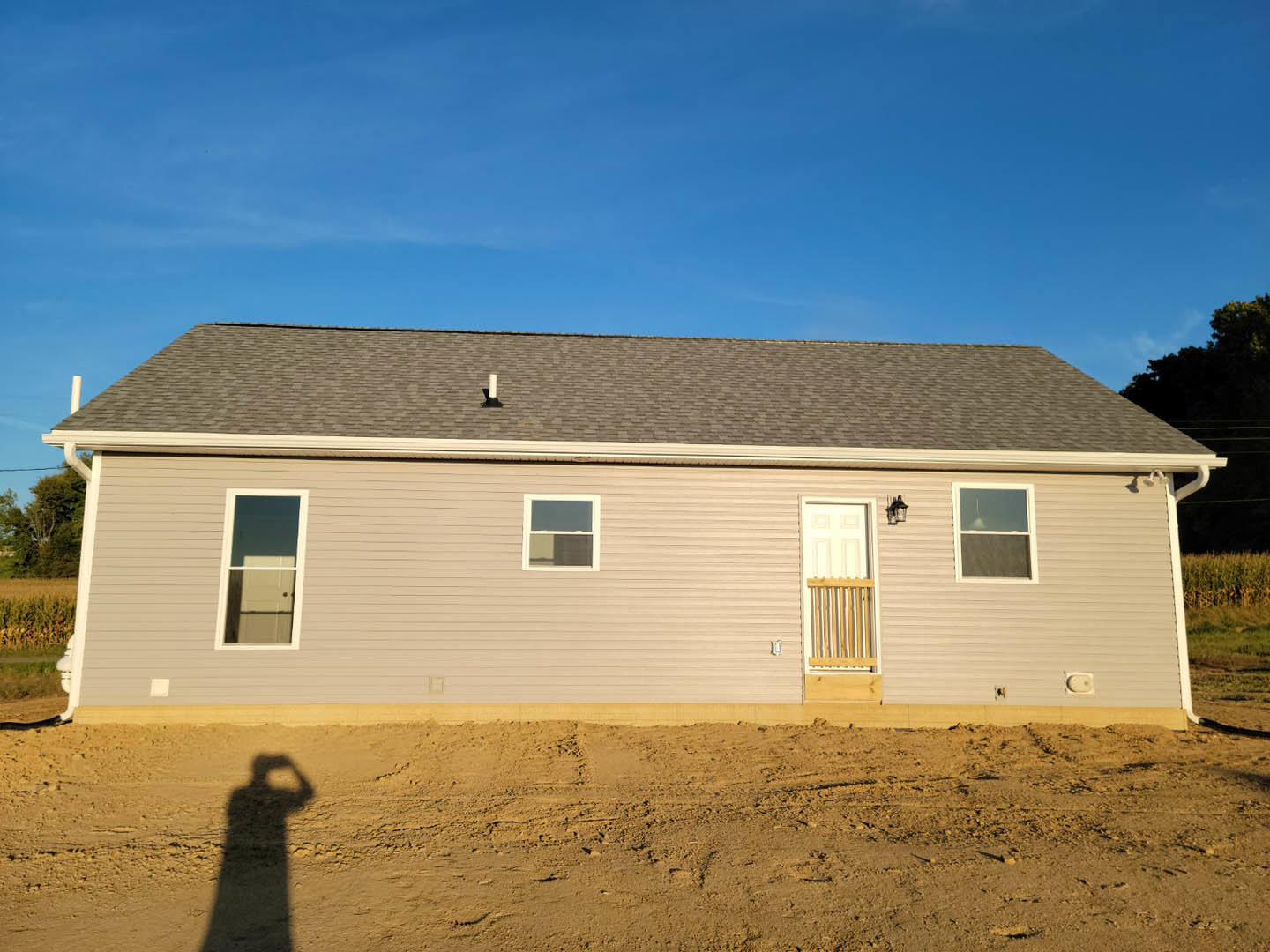 White door and window with white frame on house exterior, wooden fence in foreground, shadow of person holding object cast on siding, illuminated window, cloudy sky above
