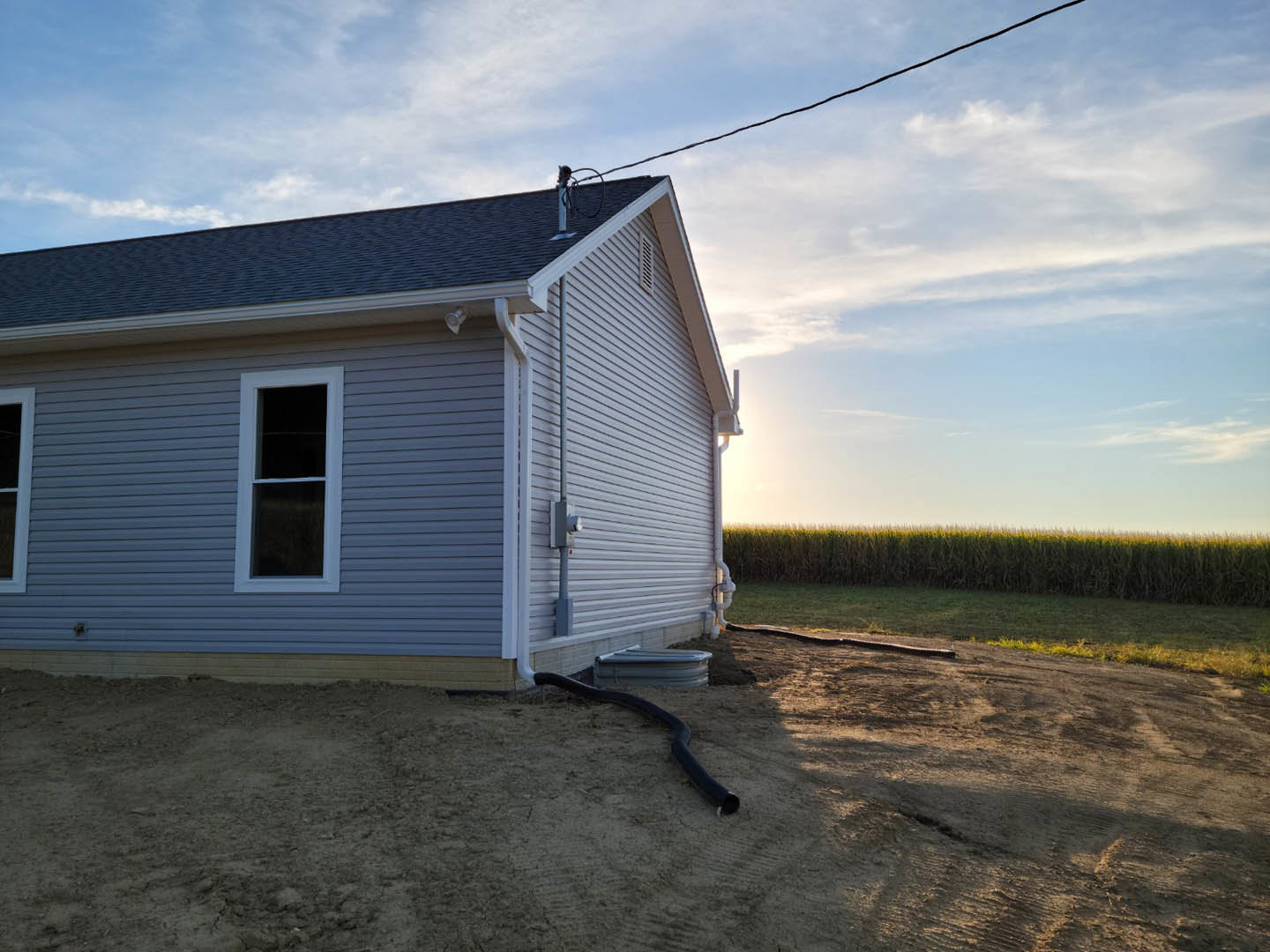 Side view of a house with white-framed window, light-colored siding, black pipe on the ground, tall grass and corn field under a partly cloudy sky