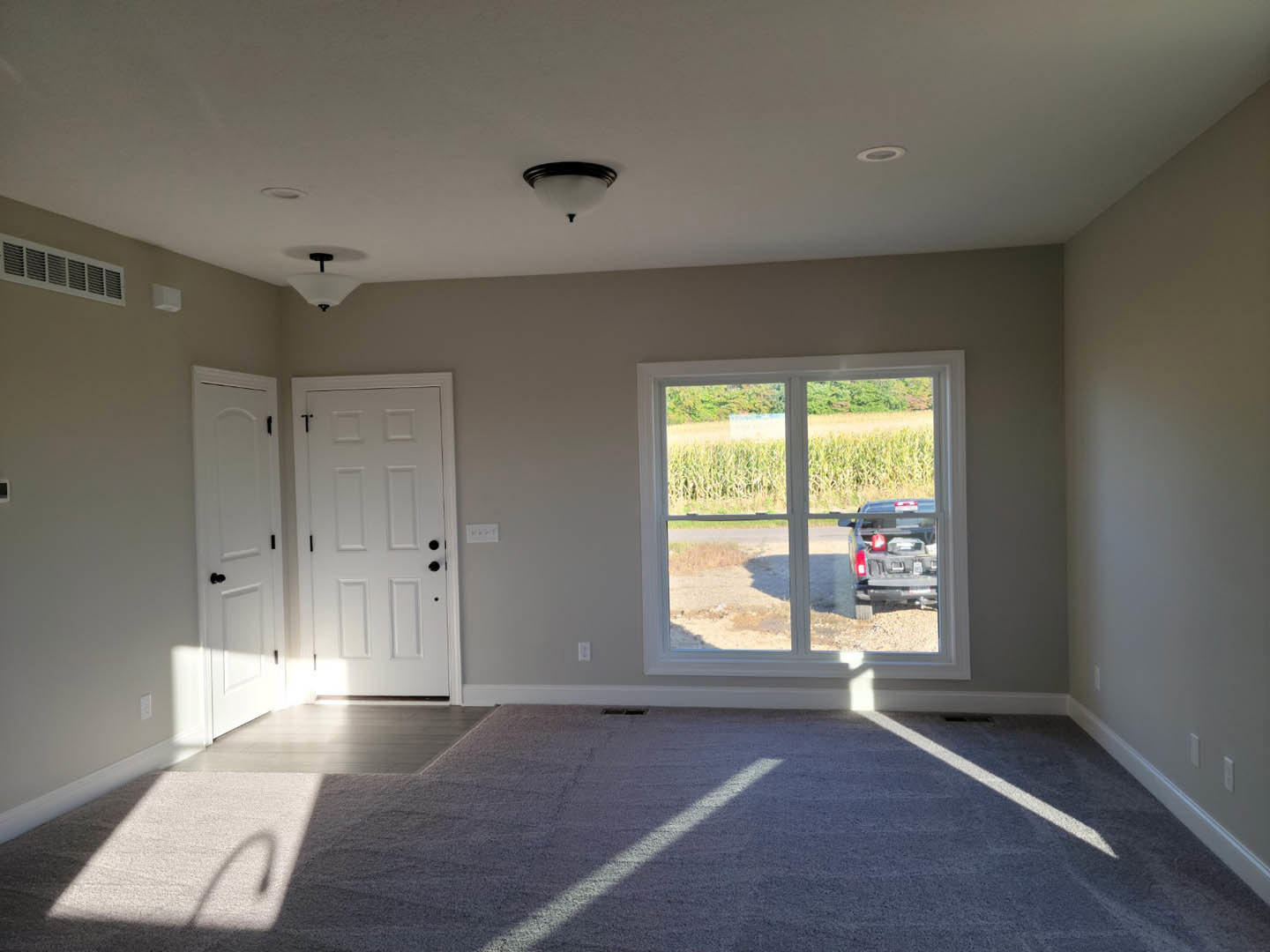 Bright room with white paneled door featuring black knobs, carpeted floor, and large window overlooking corn field and parked truck with gas can.