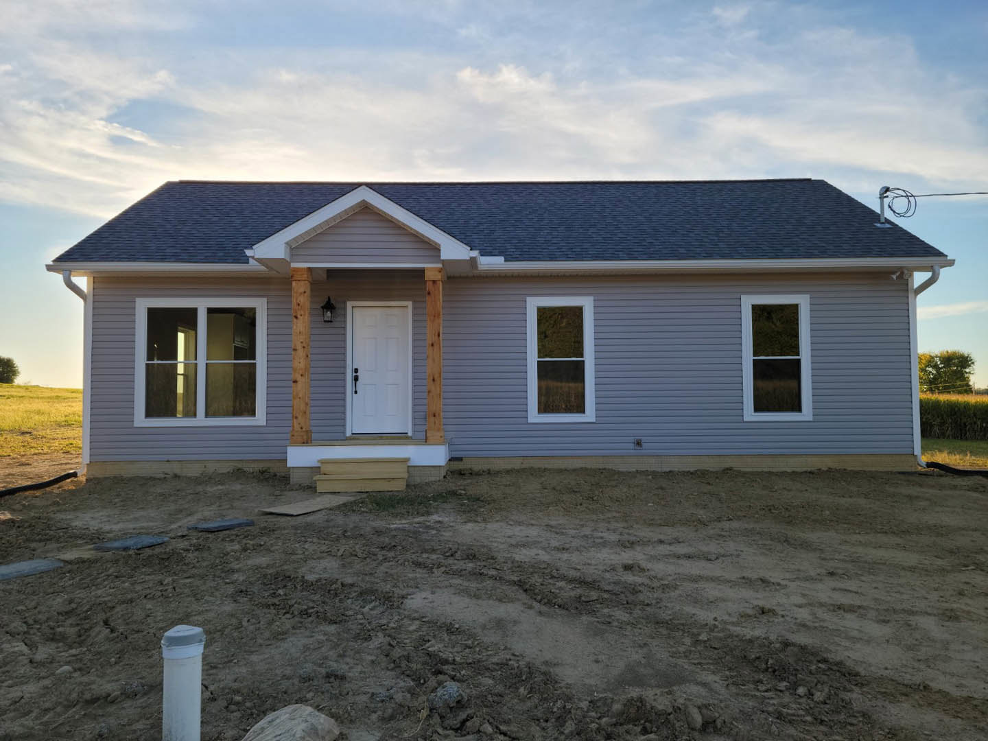 Partially built house with white siding, white door with black handle, white-framed window, exposed roof, and dirt yard in front