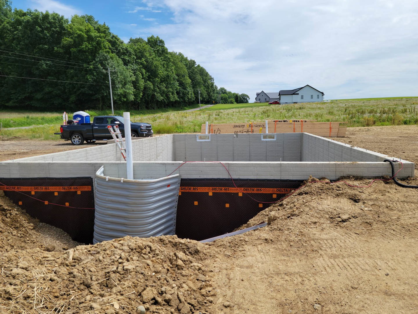 Dirt construction site with a parked truck, concrete foundation, exposed trench, water tank, power lines, and surrounding trees under blue sky