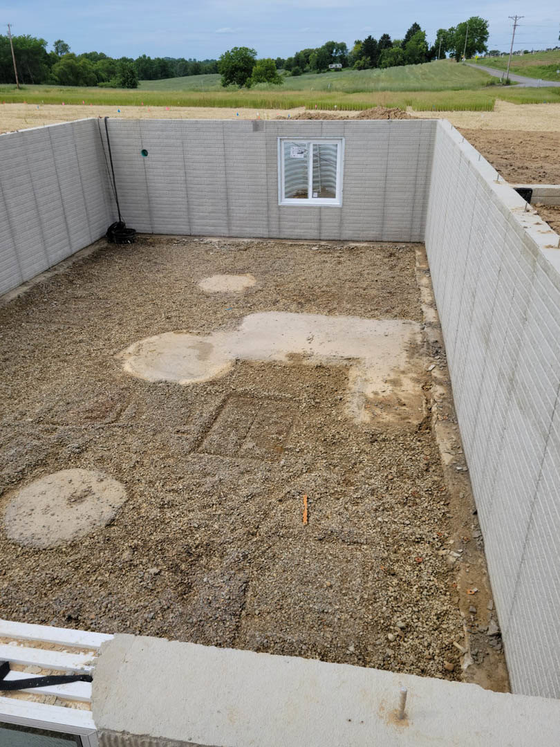 Concrete foundation with a white-framed window, dirt mound in foreground, scattered circular markings on ground, distant trees, and a white square on soil.
