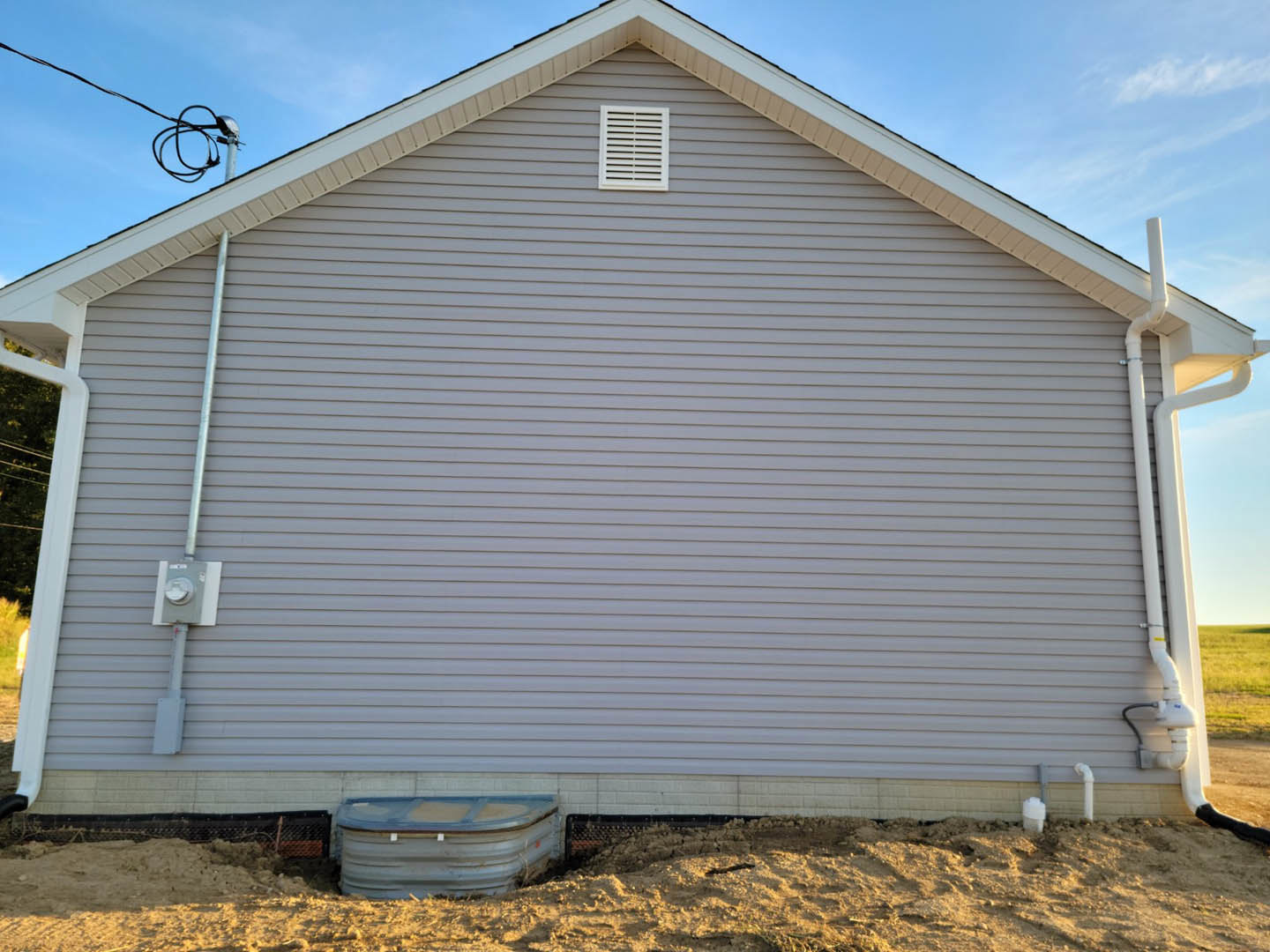 Grey house with horizontal siding, white vent on exterior wall, metal water tank beside structure, cloudy sky overhead, pile of sand in front of parked truck