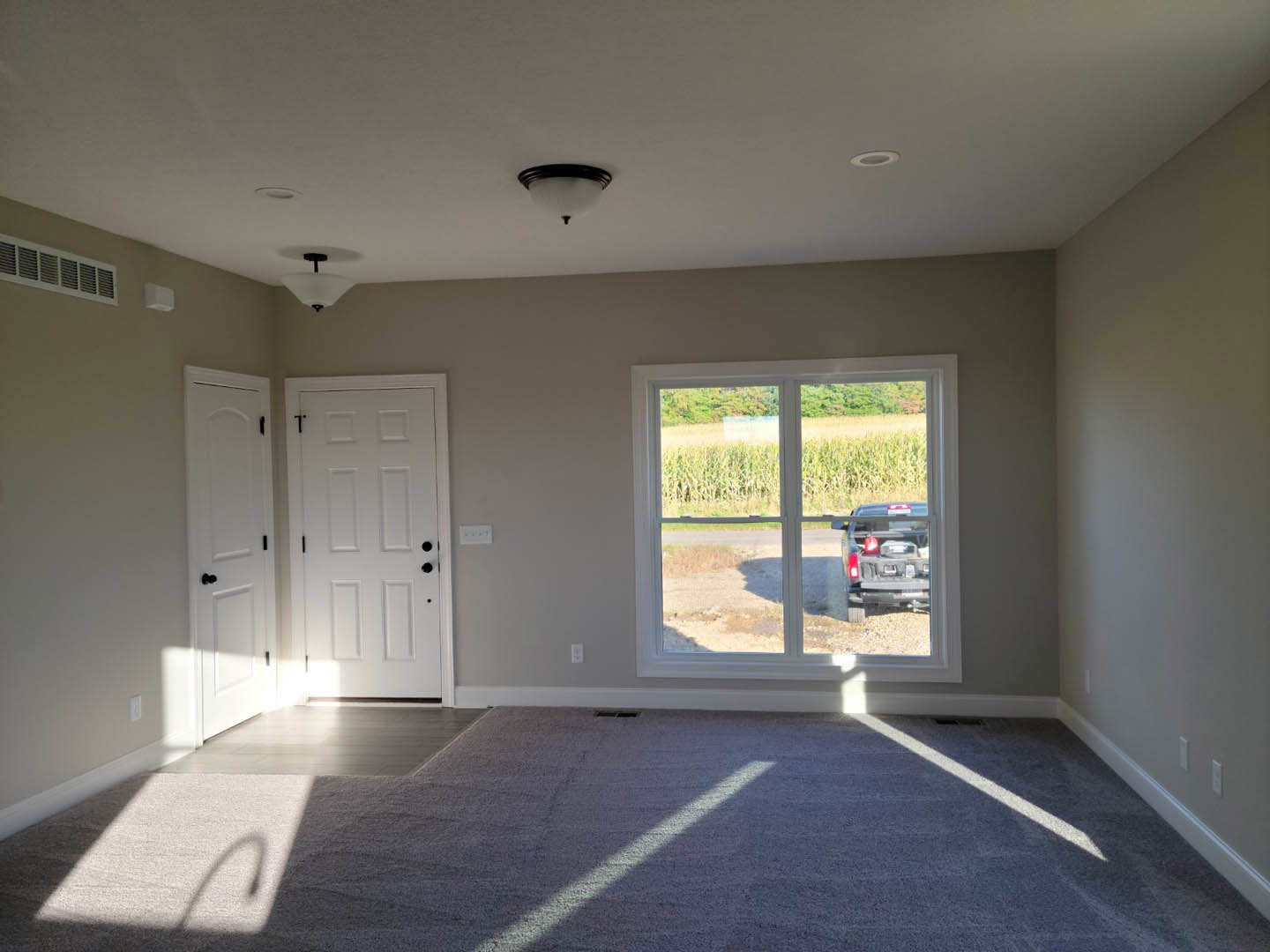 Carpeted room with white plaster walls, black-knobbed door, large window overlooking parked car and corn field, ceiling light fixture.