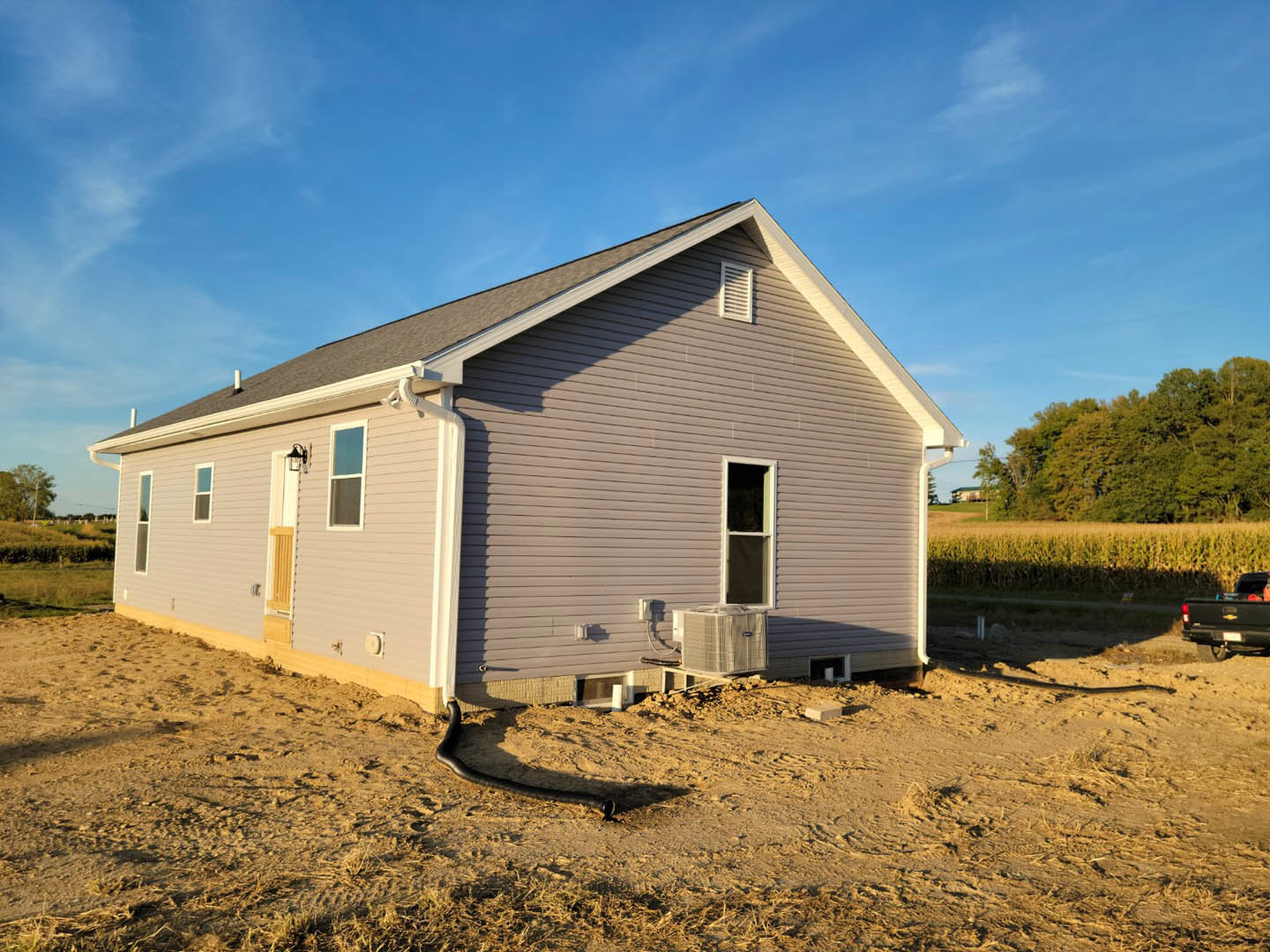 Partially built house with exposed framing and white window trim, air conditioning unit installed, black pickup truck parked nearby, surrounded by grassy field and dense trees
