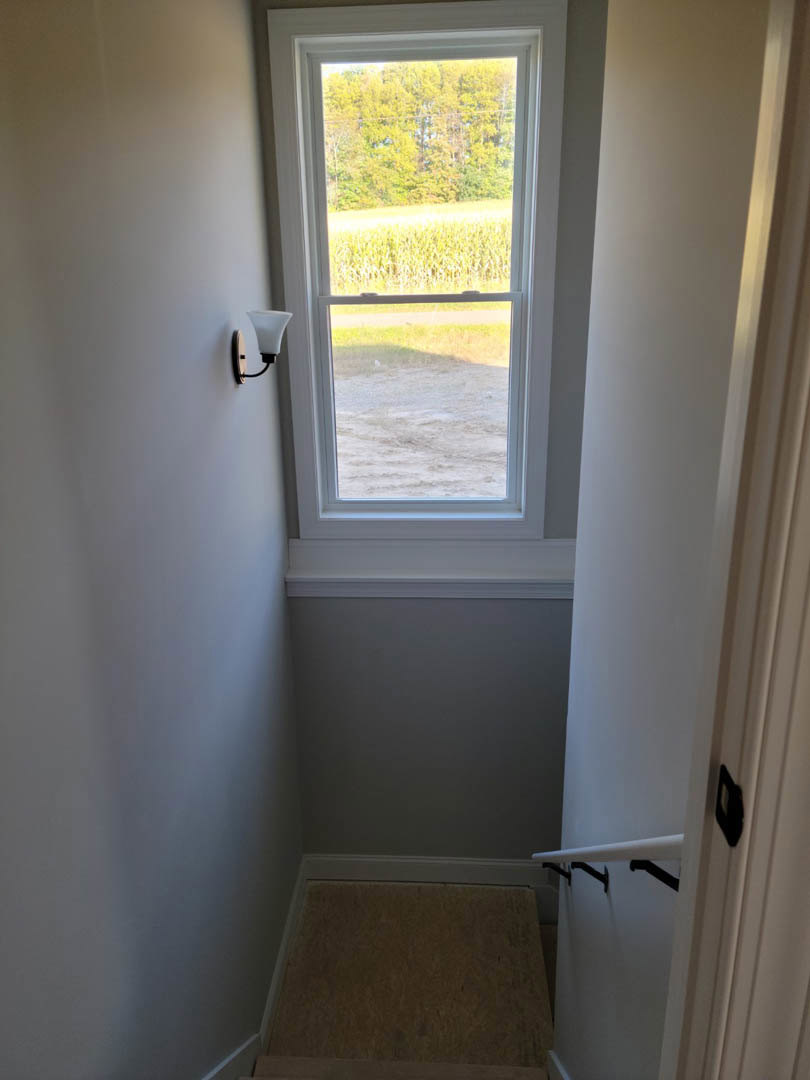 Hallway with white railing and black metal brackets, carpeted floor, large window overlooking sunlit corn field and group of trees, close-up of lamp on wall