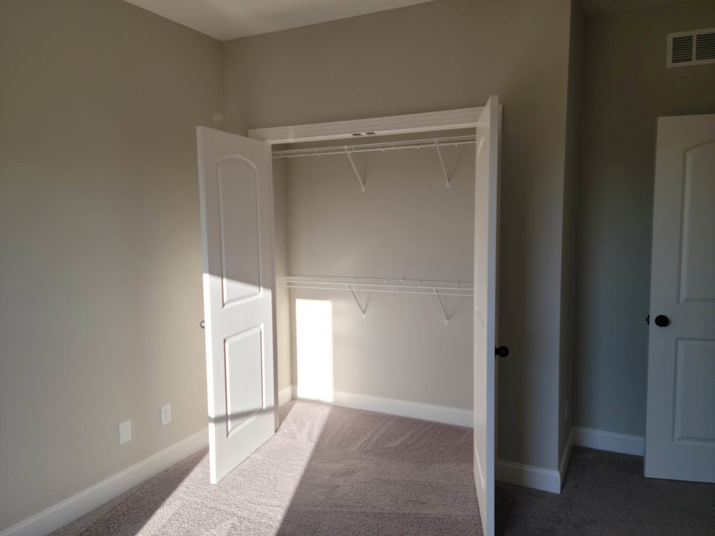 Open white door revealing closet with white carpeted floor, wall vent, and white ceiling with crown molding
