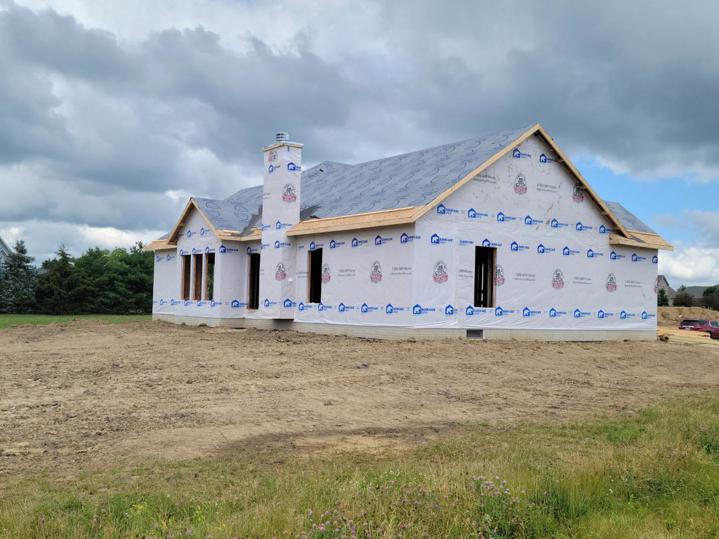 Partially built house with exposed insulation, white exterior walls, chimney, and roof featuring blue and white logo panels; surrounded by dirt field and trees under cloudy sky