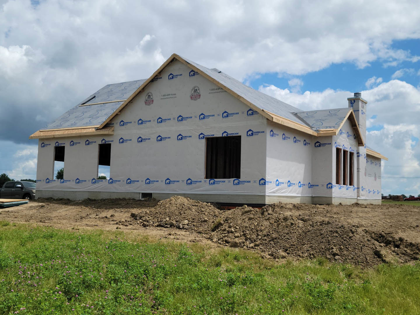 Two-story farmhouse under construction with exposed framing, dirt piles, patches of grass, and blue sky overhead