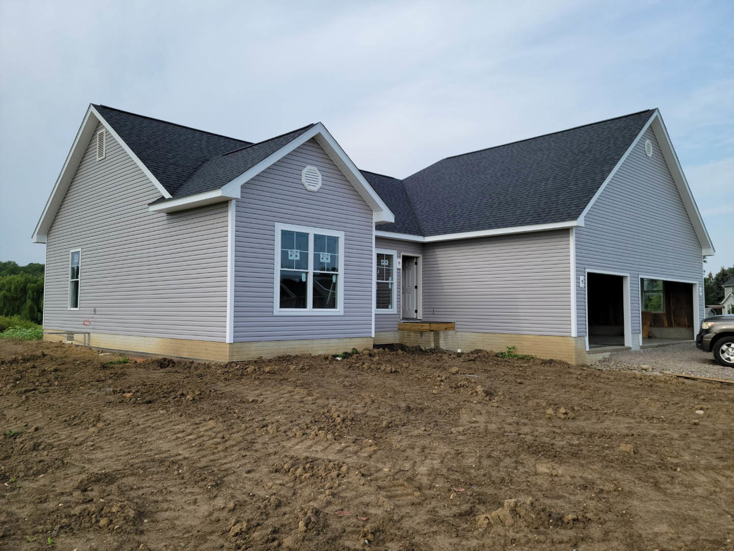 Two-story house under construction with light siding, several windows, open door revealing dark interior, dirt yard with tire tracks, and partial view of a car’s front tire