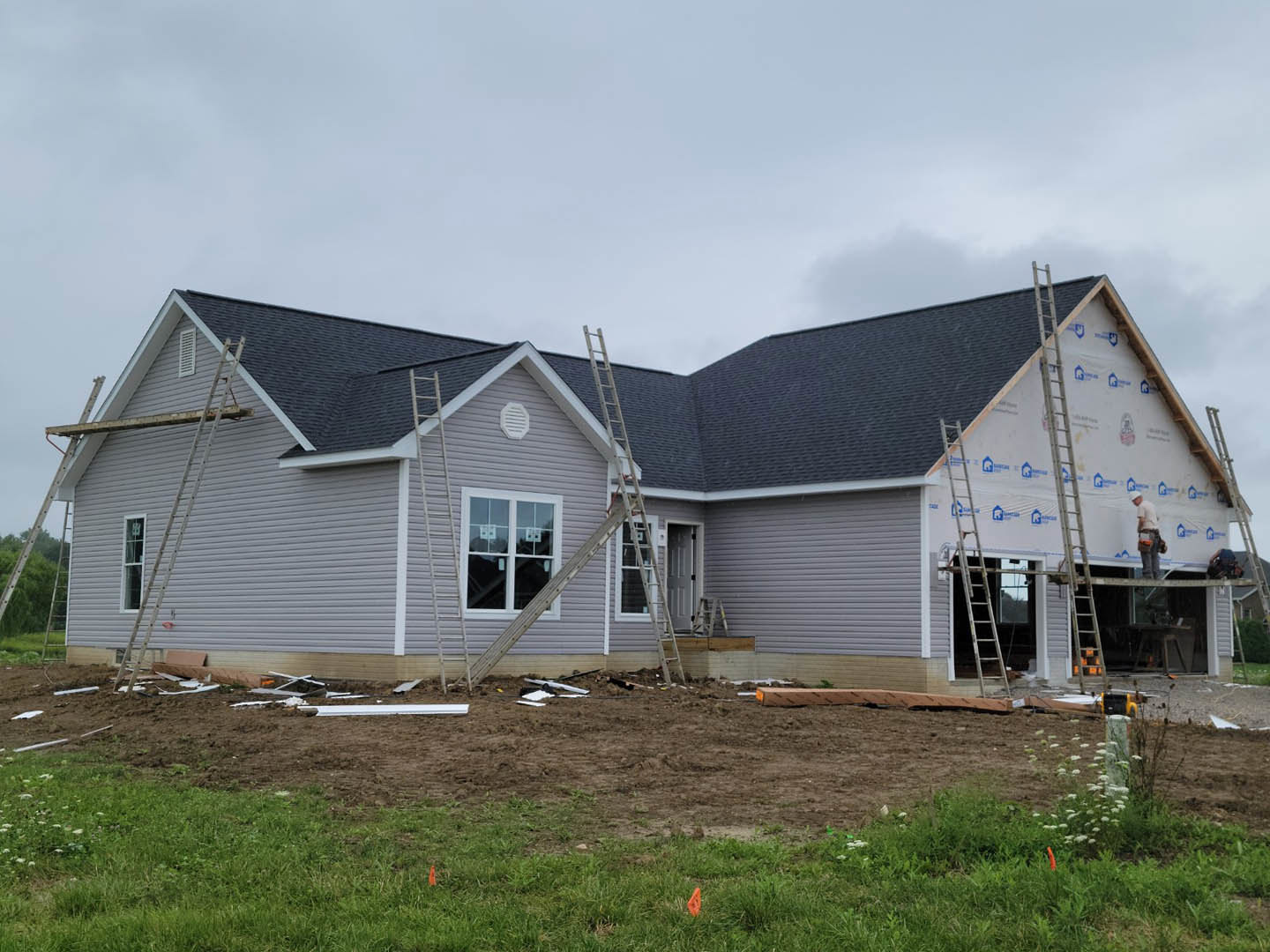 Two-story home under construction with exposed siding, multiple ladders propped against the exterior, unfinished roof, and piles of dirt and debris in the yard