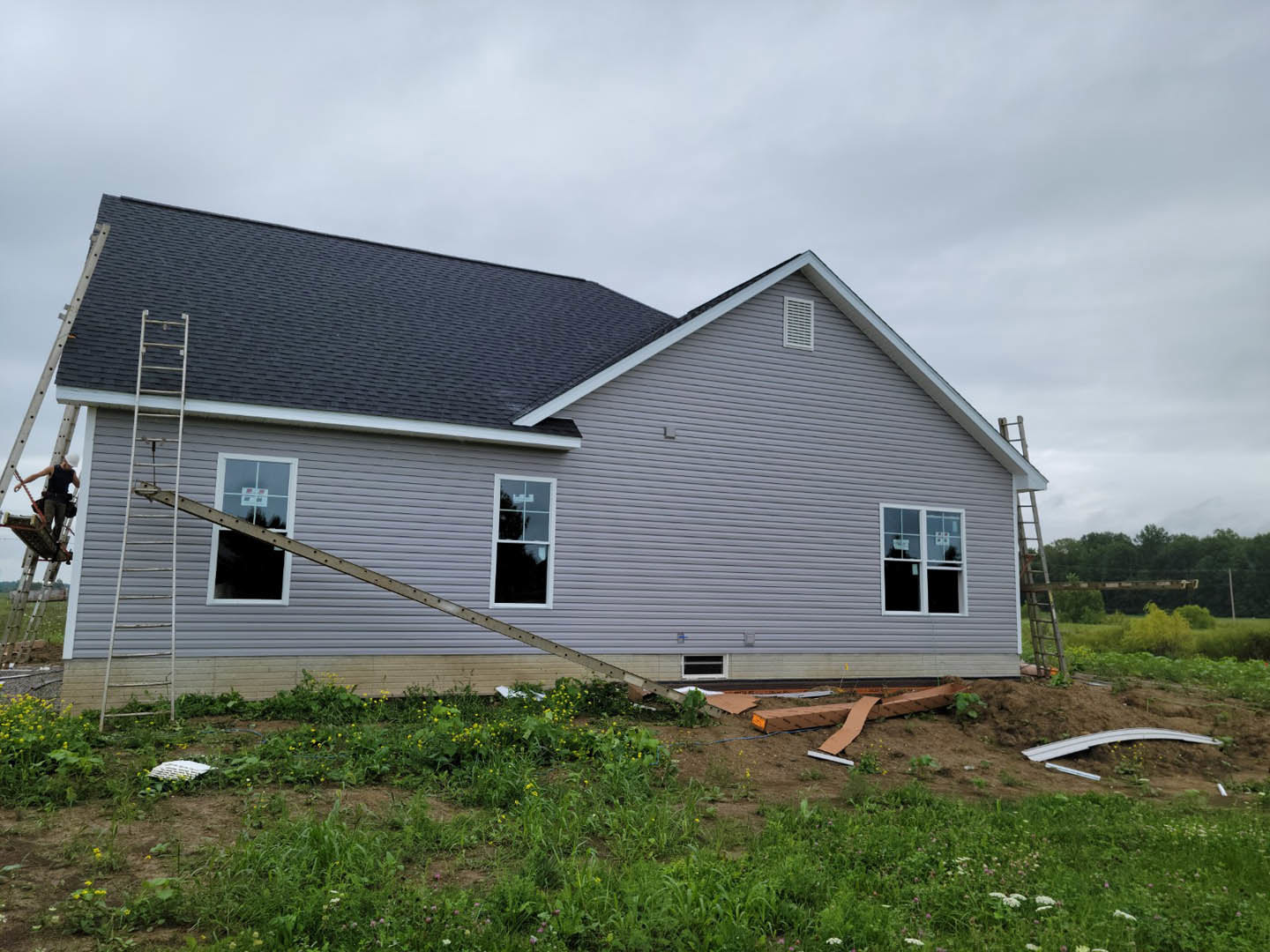 Partially constructed home with white siding, exposed window frame, ladder leaning against exterior wall, grassy yard in foreground, cloudy sky overhead