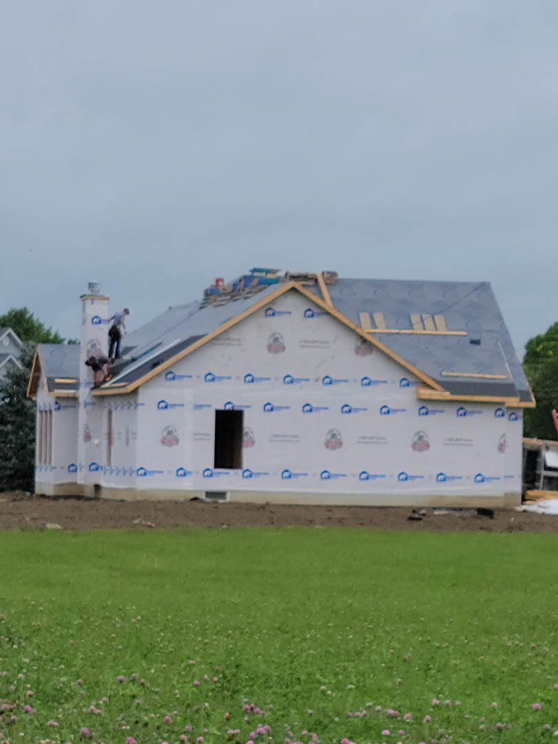 Framed house under construction with workers installing roofing, surrounded by grassy lot and scattered trees under partly cloudy sky