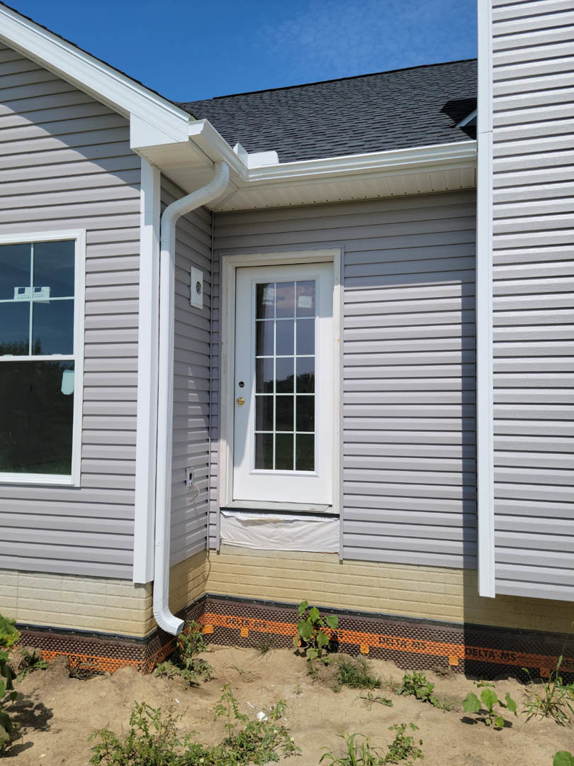 White front door with glass panes, white-framed window, light-colored siding, gutter along roof edge, small porch area, dirt landscaping in foreground