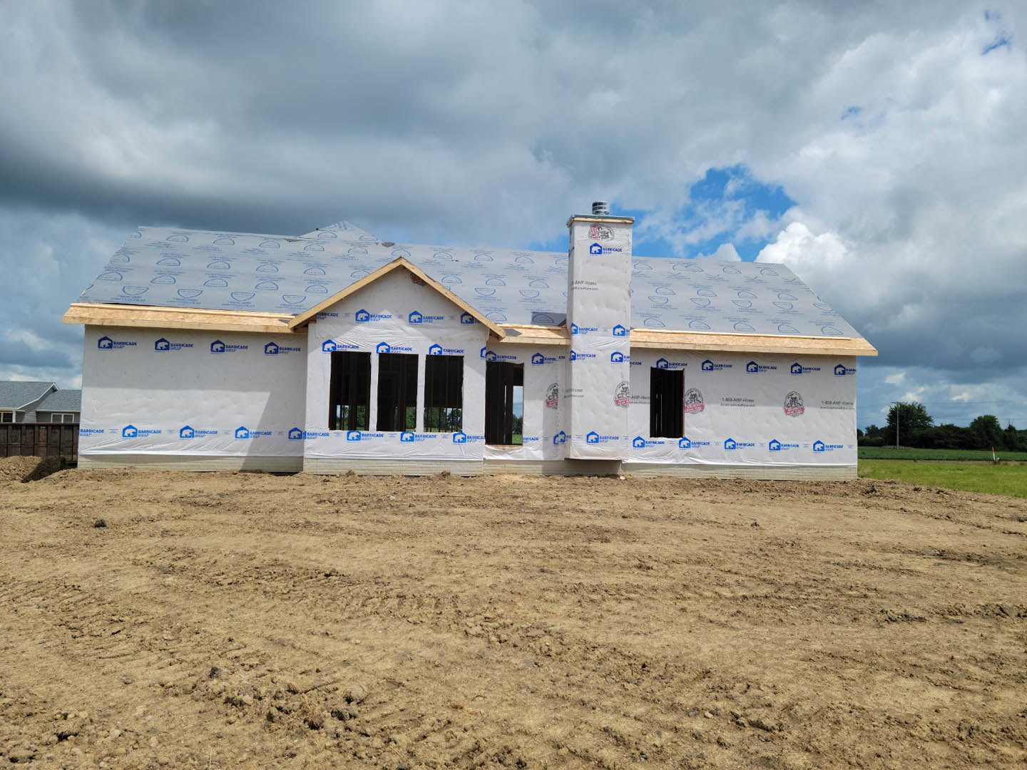 Partially built house with exposed insulation, white plastic sheets covering dirt field, stack of white boxes near foundation, cloudy sky overhead, white tarp displaying blue and
