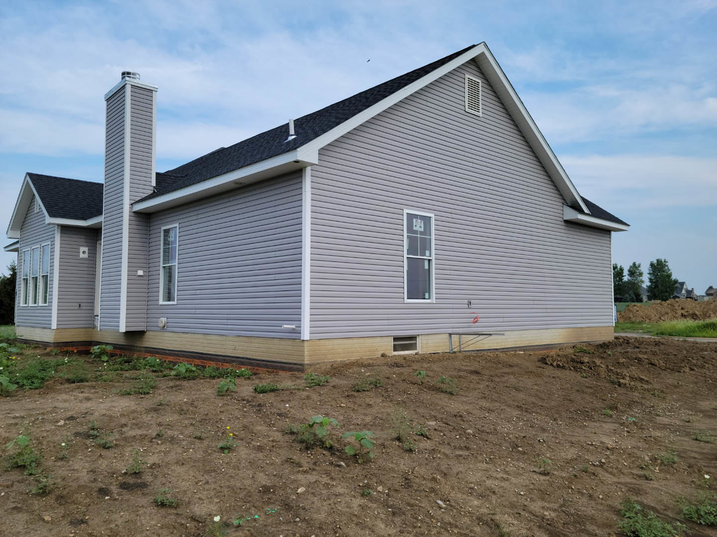 Two-story home with white-framed windows, light siding, and brick chimney, surrounded by a dirt yard with patches of grass and a leafy tree under a cloudy sky