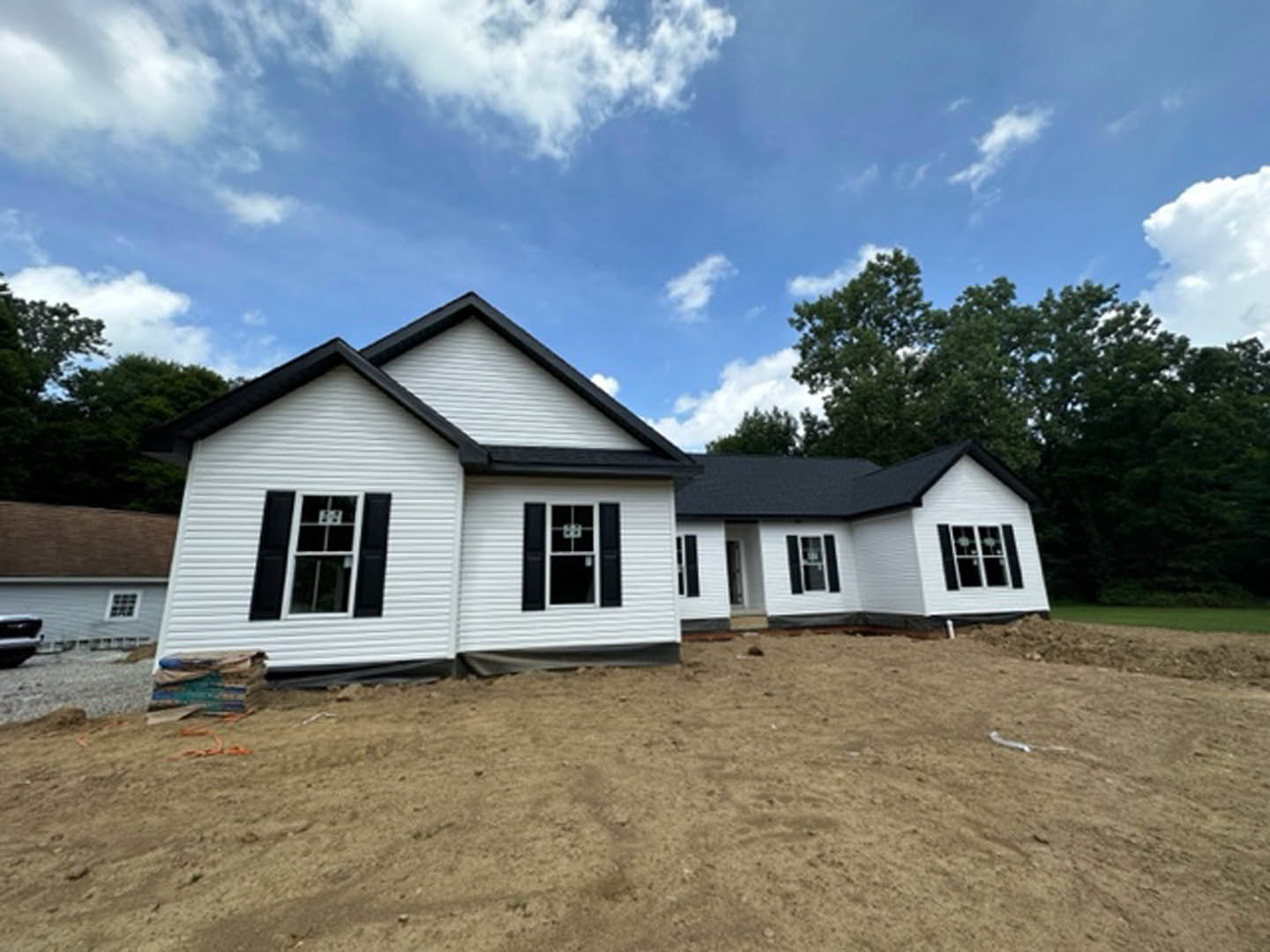 White siding house under construction on dirt lot, black roof, black shuttered windows, surrounded by trees under cloudy sky