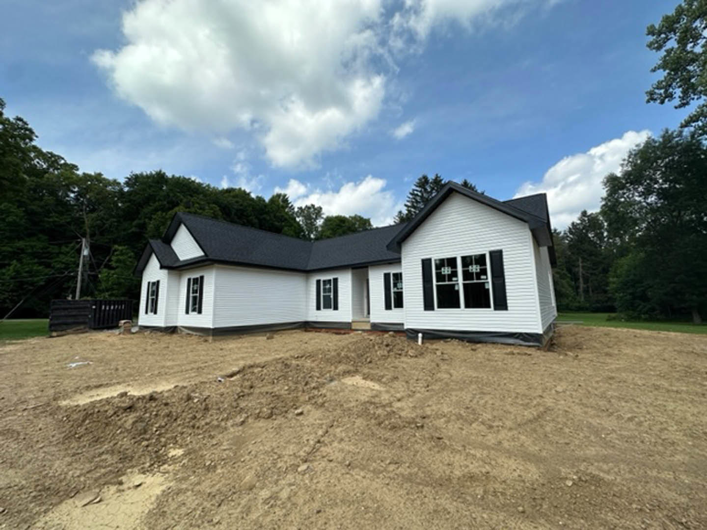 Partially built house with black roof, exposed framing, and unfinished windows, surrounded by dirt field under blue sky with scattered clouds
