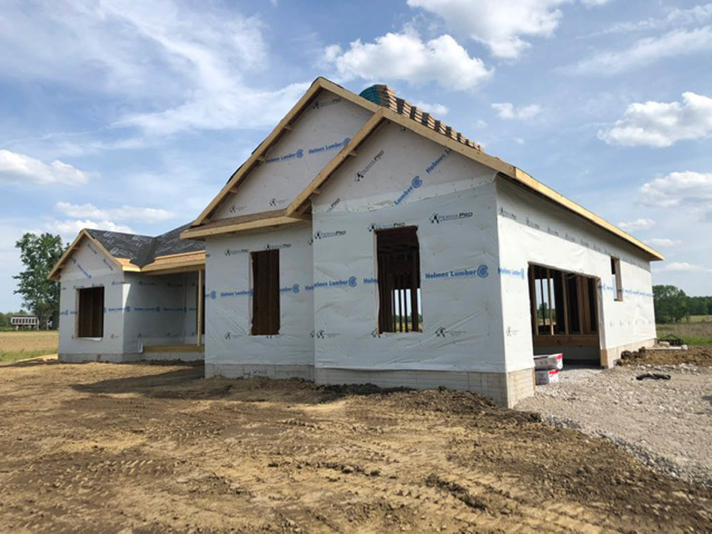 Framed custom home under construction with exposed wood, white tarp covering dirt foreground, blue sky and clouds overhead