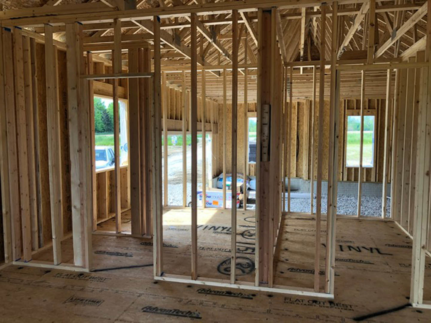 Wood-framed house under construction with exposed roof trusses, unfinished floors, and a parked car visible in the background
