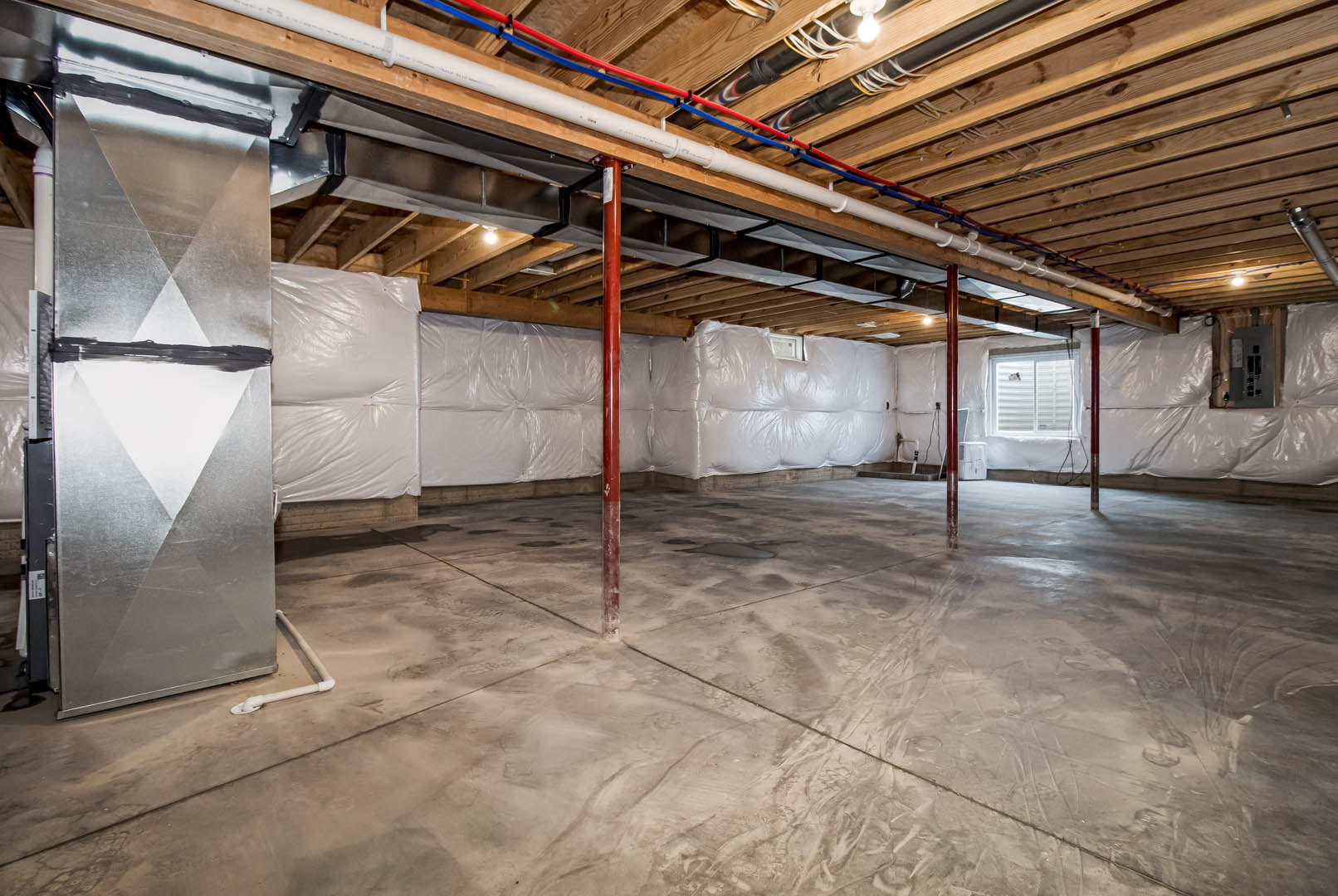 Basement with exposed metal support pole, white plastic wall covering, concrete floor, visible ceiling pipes, and building insulation.