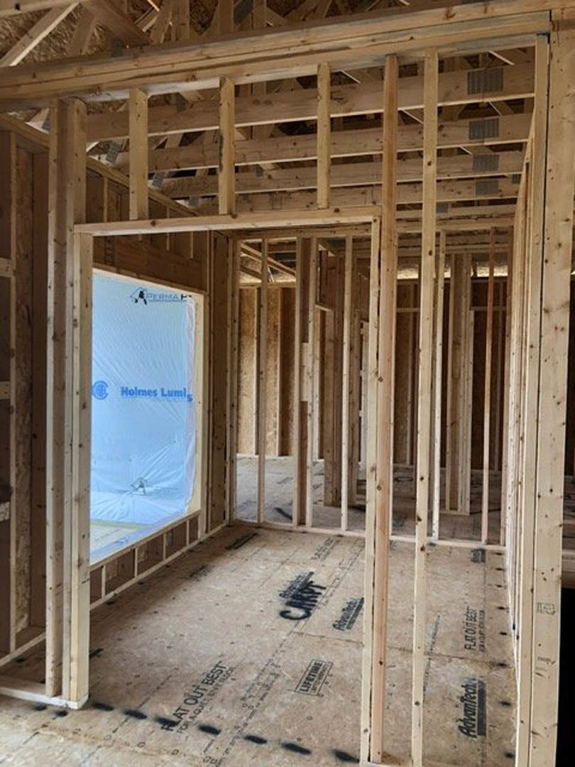 Wood-framed room under construction with exposed beams, insulation, and a window; black writing visible on framing and white surface with logo in foreground