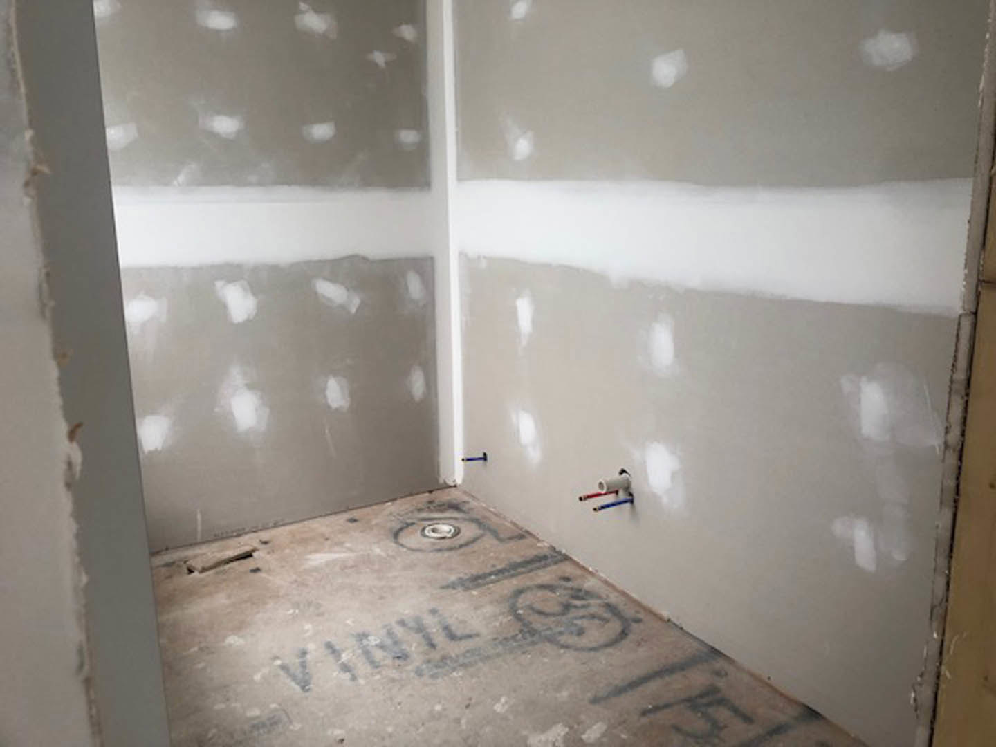 White-walled room with smooth plaster finish, concrete floor marked with black writing, exposed white pipe featuring red, blue, and white tubing.