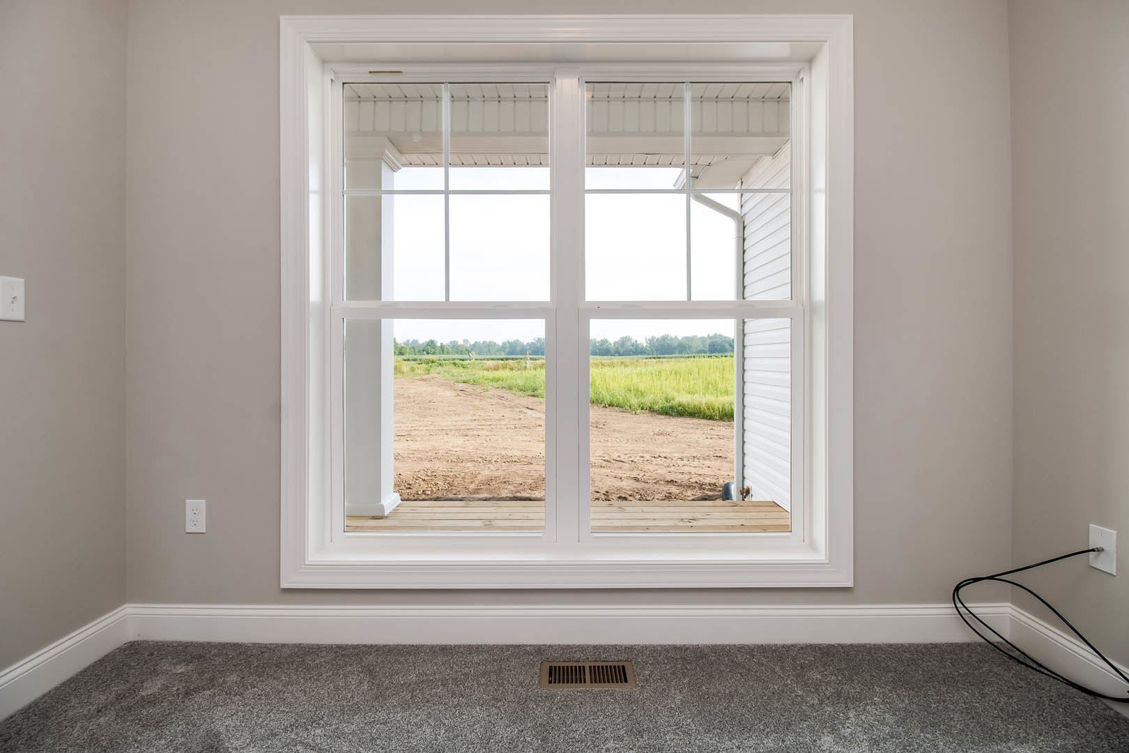 Large window with white trim and blinds, overlooking grassy field; beige carpeted floor with wall vent and white electrical outlet, black cord plugged in.
