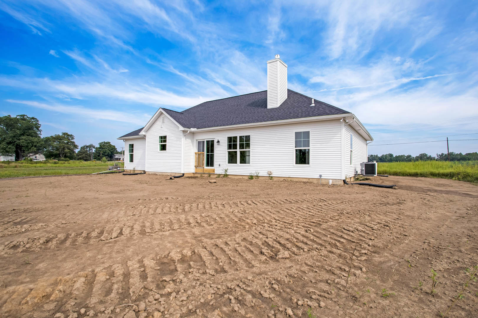 White farmhouse with prominent chimney, surrounded by dirt field with tire tracks and patches of grass, set against blue sky.