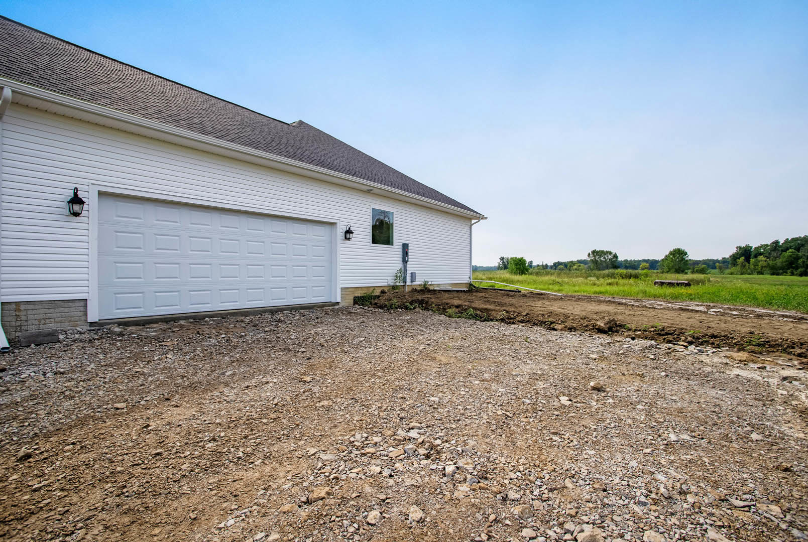 White garage with paneled door, gravel driveway bordered by patches of dirt, green-leafed tree partially visible, white siding exterior