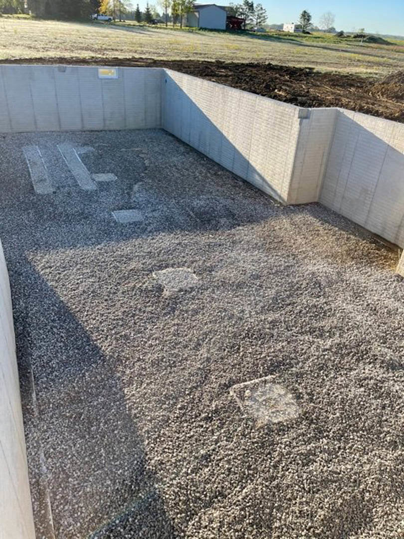 Concrete patio bordered by gravel, white exterior wall in background, concrete retaining wall with patch of dirt and small hole, sparse grass and plants along edge.