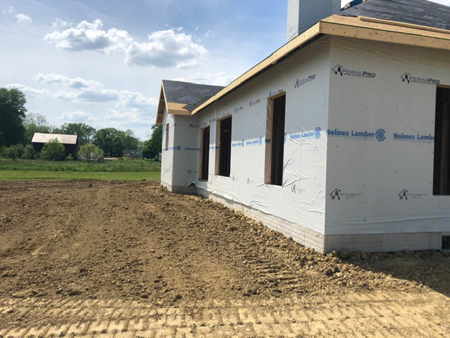 Partially built home with exposed framing, several installed windows, dirt piles surrounding the foundation, and mature trees in the background