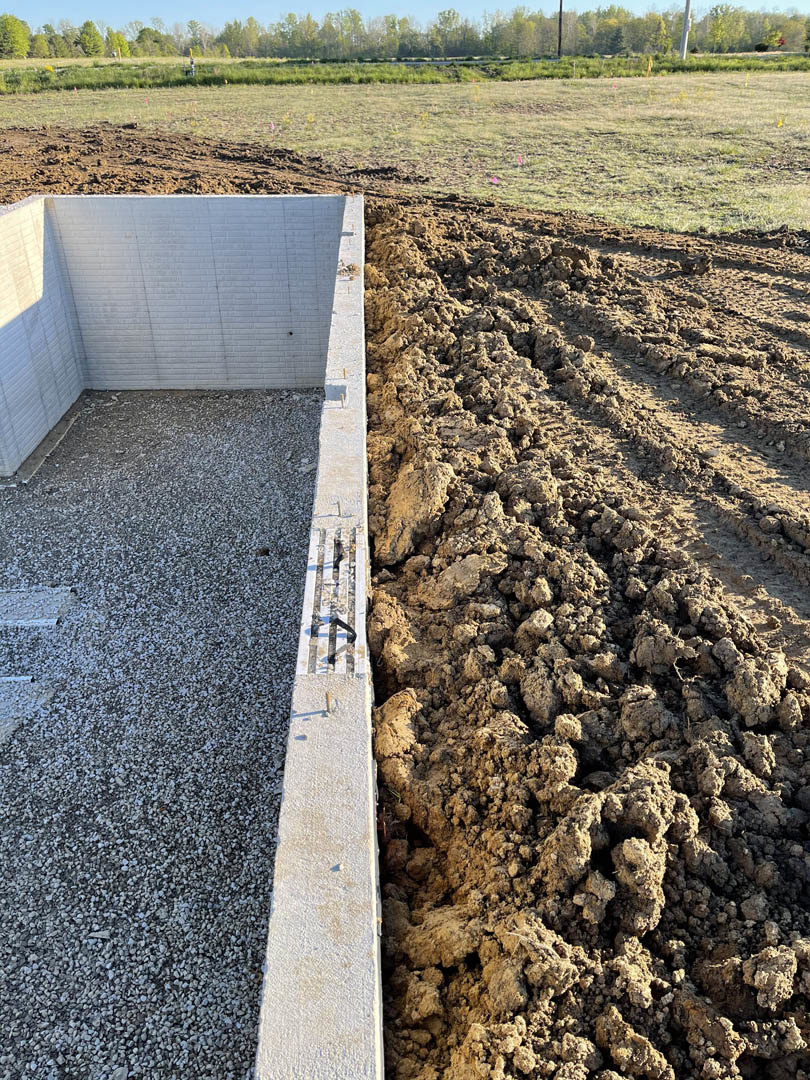 Concrete foundation bordered by a dirt field, white brick wall with black door, gravel and metal grate, dirt road with tire tracks, group of trees in the background, close-up of