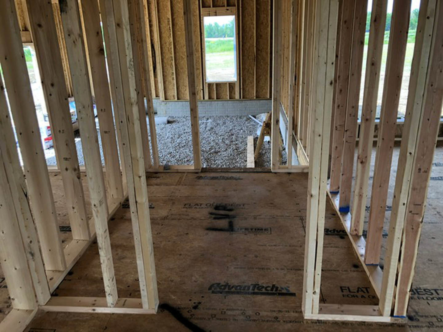 Exposed wood framing and beams inside a partially constructed home, with a window revealing grass and trees outside