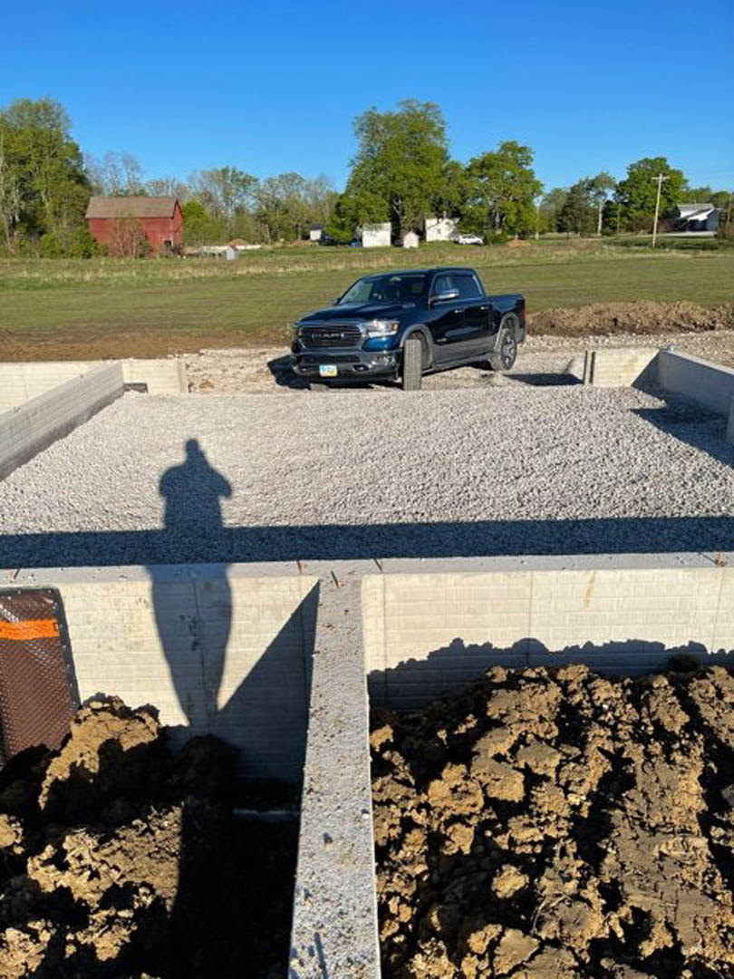 Black pickup truck parked on gravel beside unfinished concrete foundation, red building and trees in background, pile of dirt near wall.