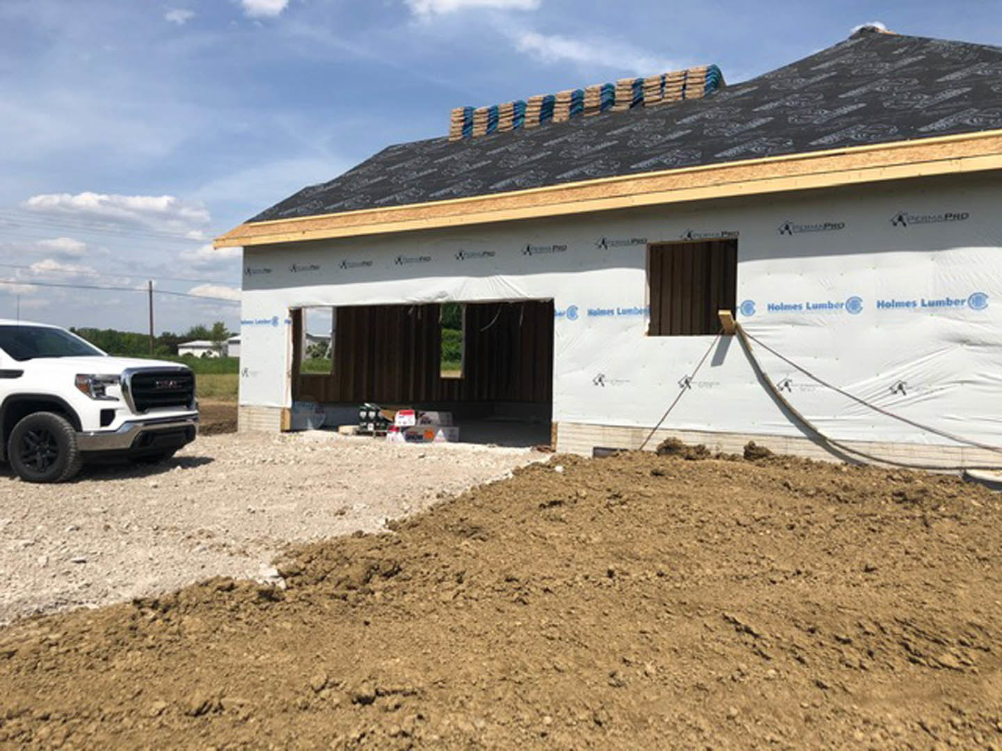 Partially built house with black roof, exposed framing, dirt mound in foreground, white pickup truck parked on gravel driveway, cloudy sky overhead