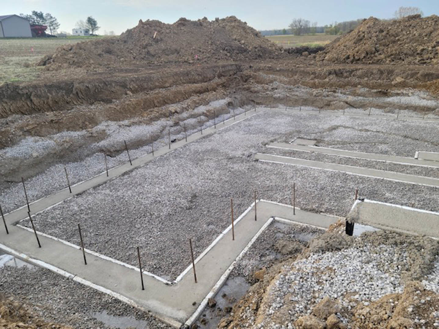 Concrete foundation with metal poles surrounded by piles of dirt and gravel, white building with flag in background, road and fence along construction site.