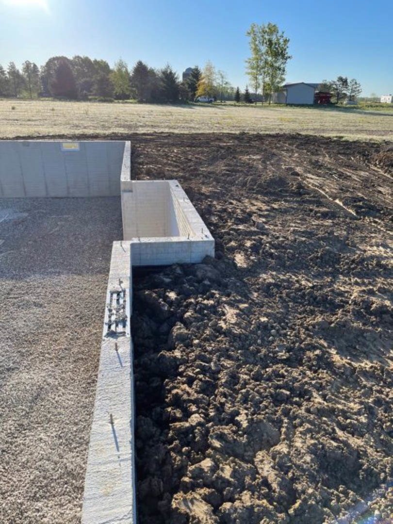 Concrete foundation slab surrounded by dirt, with green trees in the background and a white rectangular object embedded in the surface