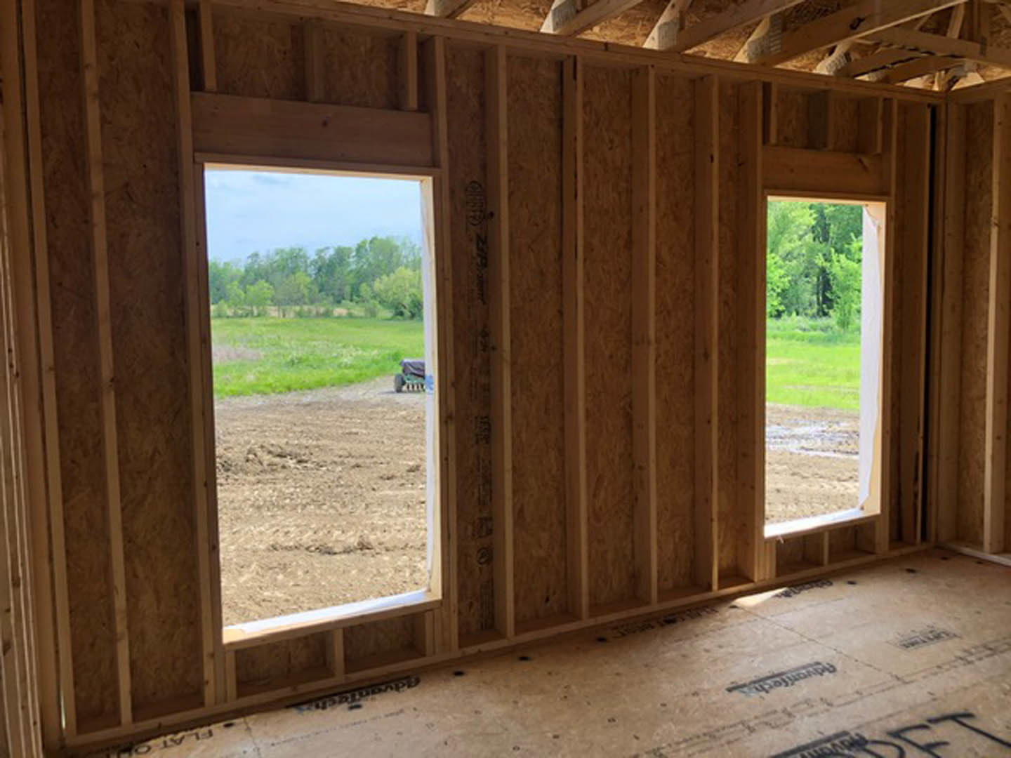 Sunlit room with two large windows, light wood flooring, and a view of grassy field through open doorway; exposed wooden ceiling beam with recessed lighting.