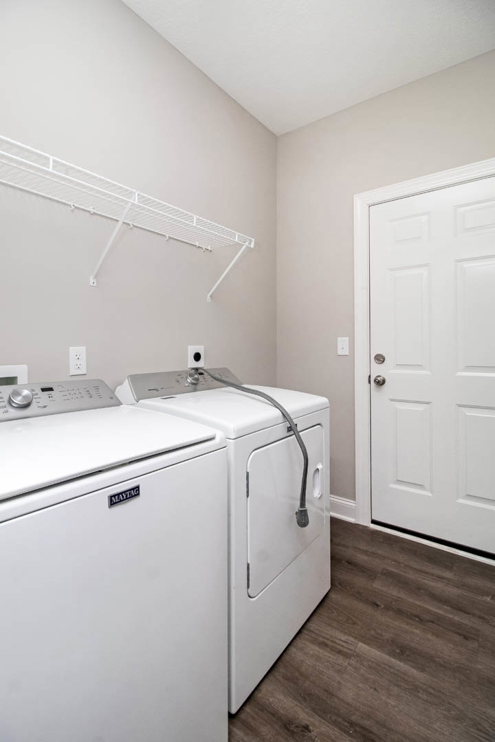 Front-loading washer and dryer set side by side in a laundry room with wood flooring, white cabinetry, and a wall-mounted white shelf