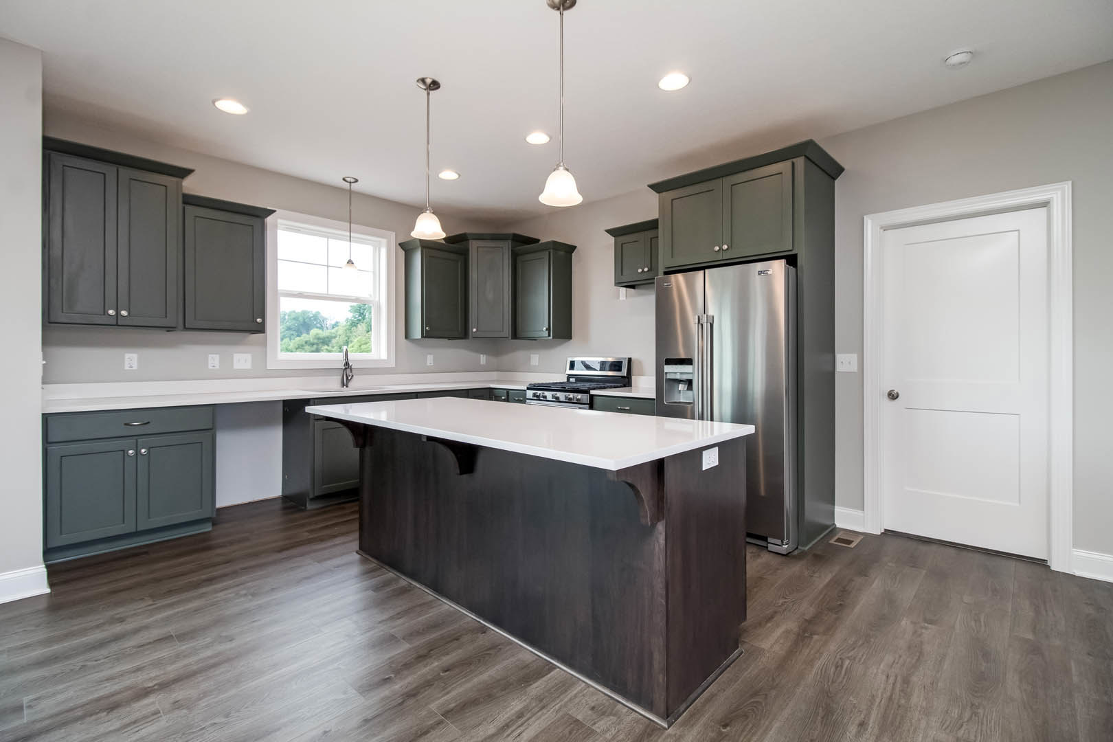 Kitchen featuring a large island with white countertop, stainless steel refrigerator, white cabinetry, silver door handle, white walls, metal support pole, and window allowing