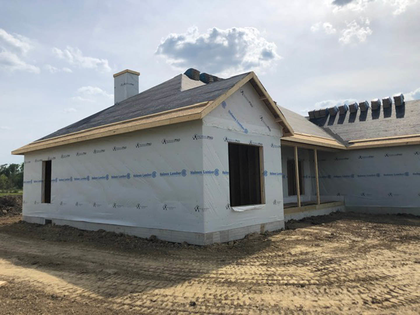 Partially built house with exposed framing, black front door, white chimney, tire tracks in dirt foreground, blue sky with scattered clouds